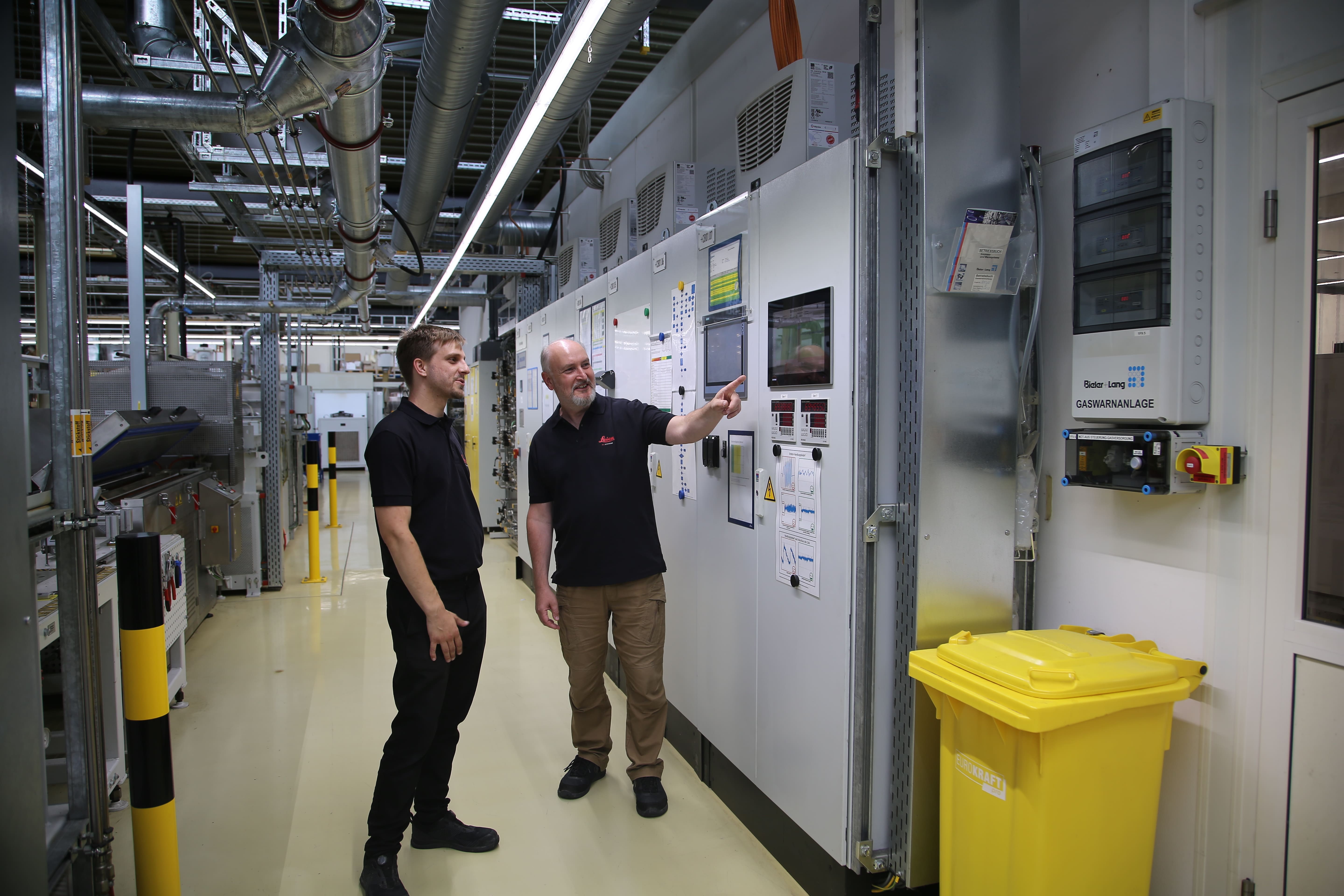 Der Geschäftsführer der Leica Biosystems Nußloch GmbH und ein Mitarbeiter sind vor deren Anlagen zu sehen.